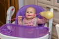 One-year-old baby sitting in a purple chair in the kitchen with a yellow ball in her teeth Royalty Free Stock Photo