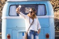 One Young Adult Woman Taking Selfie Picture With Phone Against A Blue Classic Van. Travel And Hippy Lifestyle People Concept. One