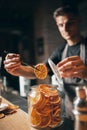 One young handsome barista in apron serves hot aromatic coffee pulling slices of dried orange from jar