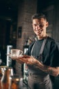 One young handsome smiling barista in apron serves hot aromatic coffee, cappuchino of latte in a white cup