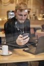 One young man, sitting in coffee shop and using his smart-phone, with laptop and cup nearby. Shoot thought window outside with Royalty Free Stock Photo