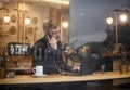 One young man, 20-29 years, sitting in coffee shop and using, his laptop while talking over phone. Shoot thought window outside Royalty Free Stock Photo