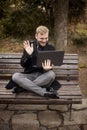 One young relaxed and smiling man, sitting casually on bench with crossed legs in public park, using laptop with video call Royalty Free Stock Photo