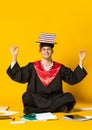 Portrait of young smiling man, student in graduation cap and gown sitting on floor witth books on head isolated over Royalty Free Stock Photo