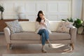 Calm young woman houseowner sitting on couch using mobile phone