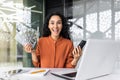 Online casino. Young muslim woman sitting in office at table with laptop, holding cash in hand and phone, playing online