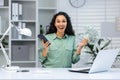 Online casino. Young muslim woman sitting in office at table with laptop, holding cash in hand and phone, playing online