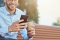 Online chatting. Side view of a happy businessman using smartphone and drinking coffee while relaxing on the bench Royalty Free Stock Photo