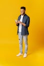 Online communication. Full length portrait of indian man using smartphone standing isolated on yellow background