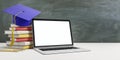 Online education concept with stack of books, blue graduate cap and blank white laptop screen on wooden table at blackboard