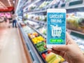 Online grocery shopping concept: Woman hand holding smart phone for ordering food onscreen with icon media on supermarket