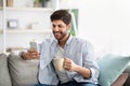 Online life concept. Positive arab man holding modern smartphone and cup of coffee at home, free space