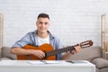 Online music lessons. Cheerful guy with guitar sits on couch in living room interior and records video or makes record