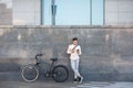 Online outdoors. Businessman with bicycle and cup of coffee, looks at smartphone leaning against the wall