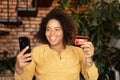 Young happy african-American woman holds bank card and smartphone sitting on the armchair Royalty Free Stock Photo