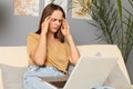 Online research. Busy programming. Tired brown haired woman wearing beige T-shirt and jeans sitting on sofa and working on laptop