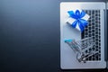 Online shopping background. Laptop computer, shopping trolley and white gift with blue ribbon on dark. Internet purchase Royalty Free Stock Photo