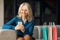 Online shopping concept. Cheerful mature woman shopaholic purchasing online, sitting on sofa near colorful shopping bags Royalty Free Stock Photo