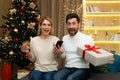 Online shopping. Happy young couple at home near Christmas tree, holding credit card, phone and gift Royalty Free Stock Photo