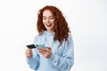 Online shopping. Portrait of redhead woman paying in internet, holding plastic credit card and smartphone, standing