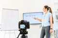 Female teacher is recording classes on a camera, she stands near flip chart and screen with a marker in hand, camera in foreground Royalty Free Stock Photo