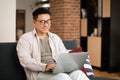 Online work. Happy asian man using laptop, typing and surfing internet sitting on sofa at home, free space Royalty Free Stock Photo