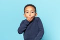 Oops. Portrait of amazed african american little boy covering mouth with hand and looking at camera, blue studio wall