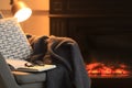 Open book, glasses and plaid on armchair near fireplace at home, space for text. Cozy atmosphere Royalty Free Stock Photo