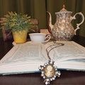 open book, necklace and medallion as a bookmark.  in the background a teapot, a tea cup, flowers and old books Royalty Free Stock Photo