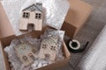 Open box with decorative house figures, adhesive tape, foam peanuts and bubble wrap on wooden table