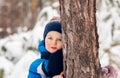 Open close-up portrait of cute happy smiling boy in blue jacket and knitted hat looking behind the tree Royalty Free Stock Photo