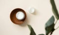 Open cream moisturizer jar in wooden plate on light beige background with blur eucalyptus leaf, above. Set natural organic eco