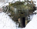 Open dark cellar door in sandstone rock cave with moss and lichens and long hanging icicles, white snow background.