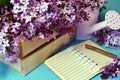 Open diary note book with lilac flowers in wooden box and watering can over blue background
