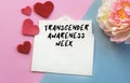 An open envelope with the text TRANSGENDER AWARENESS WEEK, on a pink and blue background with a decor of felt hearts and a flower.