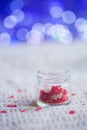 Open little glass jar filled with small sweet candies in form of heart on white knitted fabric with blue bokeh background