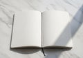 An open notebook with blank pages lies on a smooth, white marble surface. Sunlight casts a diagonal Royalty Free Stock Photo