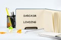 An open notebook displays the words Sarcasm Loading in bold letters. A pen holder filled with writing tools and crumpled paper sit Royalty Free Stock Photo