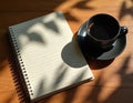 Open notebook with handwritten text sits beside black coffee cup on wooden table. Leaf shadows create dappled light pattern on Royalty Free Stock Photo