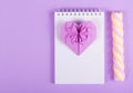 Open notebook, Valentine origami and marshmallow stick on a purple background.