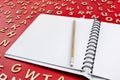 An open notebook with white pages and a wooden pencil lying on a red background with scattered letters made of wood.