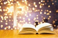 Open old bible on a wood table with blurred cross & colourful sparking lights as background