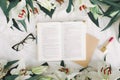 Opened book, red lipstick, glasses and fresh flowers on the white bed. Flat lay, top view Royalty Free Stock Photo