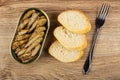 Opened jar with canned sprats, slices of bread, fork on wooden table. Top view
