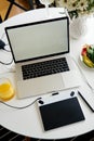 Opened laptop, graphics tablet and breakfast with juice on white table, empty screen for mockup design.