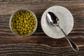 Opened jar with green peas, spoon in bowl on wooden table. Top view