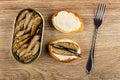 Opened jar with sprats, slice of bread with mayonnaise, sandwich with sprats, fork on wooden table. Top view