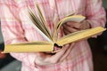Opened thick, old book of yellow color in female hands, against the background of a pink plaid shirt.