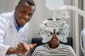 Optician Doing Optometry Eye Exam For Black African teen girl Patient. Male optometrist with phoropter while examining patient