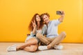 Optimistic couple young man and woman 20s sitting on floor together and showing peace sign while taking selfie on smartphone, iso Royalty Free Stock Photo